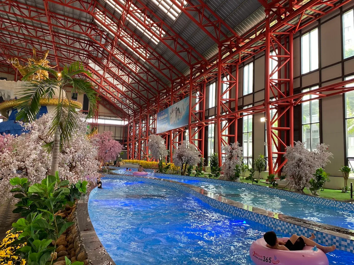 Không gian công viên nước trong nhà với dòng sông lười, hồ bơi và cảnh quan hoa anh đào trang trí.