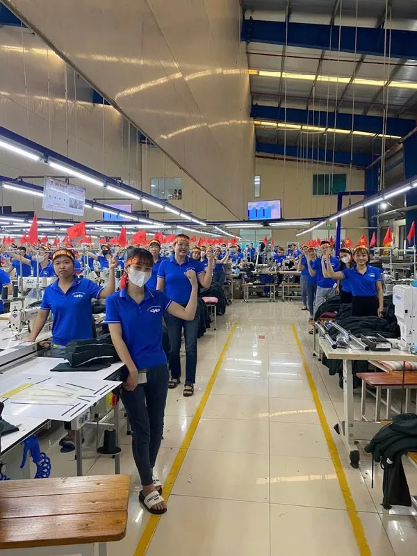 Không gian xưởng may với nhiều công nhân mặc áo xanh và các dãy máy may.