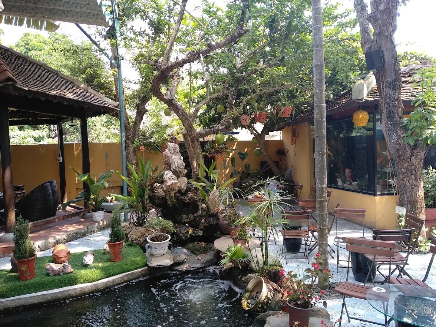 Không gian sân vườn của quán cà phê với bàn ghế ngoài trời, cây xanh và hồ cá cảnh.