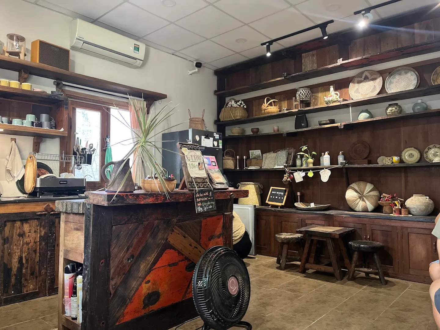 Không gian quán cà phê mộc mạc với quầy gỗ, kệ đồ thủ công và khu vực ghế ngồi.