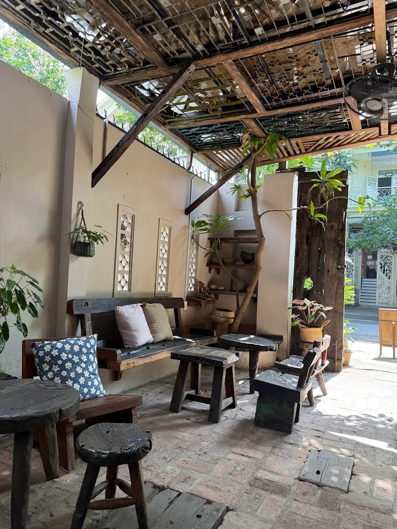 Không gian sân cà phê mộc mạc với bàn ghế gỗ, cây xanh và mái che độc đáo.