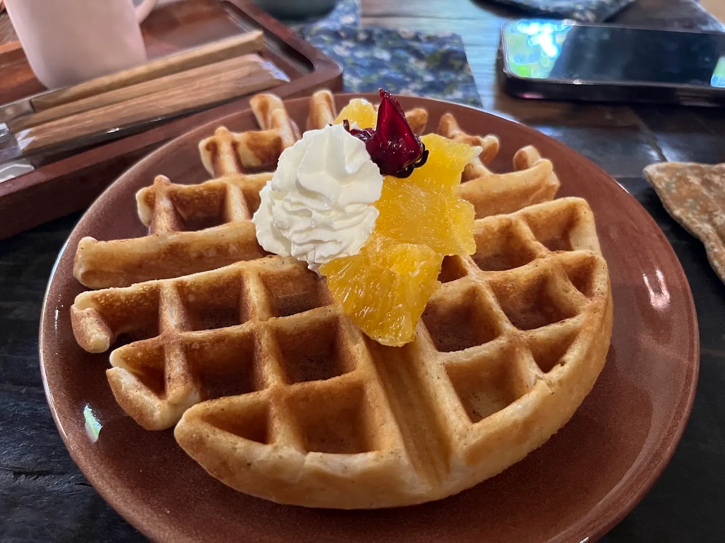 Bánh waffle phủ kem tươi, lát cam và cánh hoa khô trên đĩa.