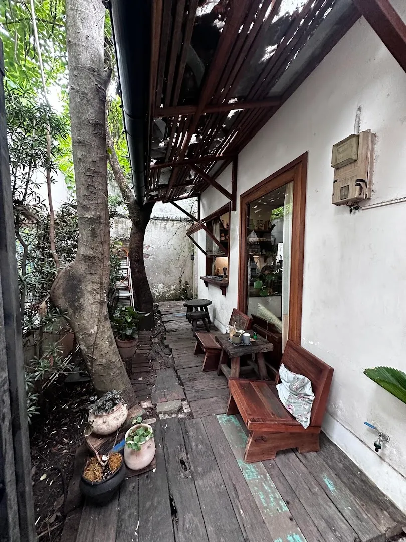 Không gian ngồi ngoài trời mộc mạc với bàn ghế gỗ, cây xanh và lối đi lát ván.