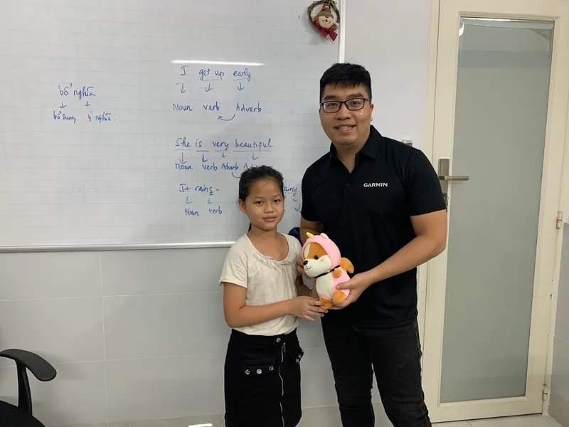 Không gian lớp học với bảng trắng ghi chú ngữ pháp tiếng Anh và hai người đứng phía trước.