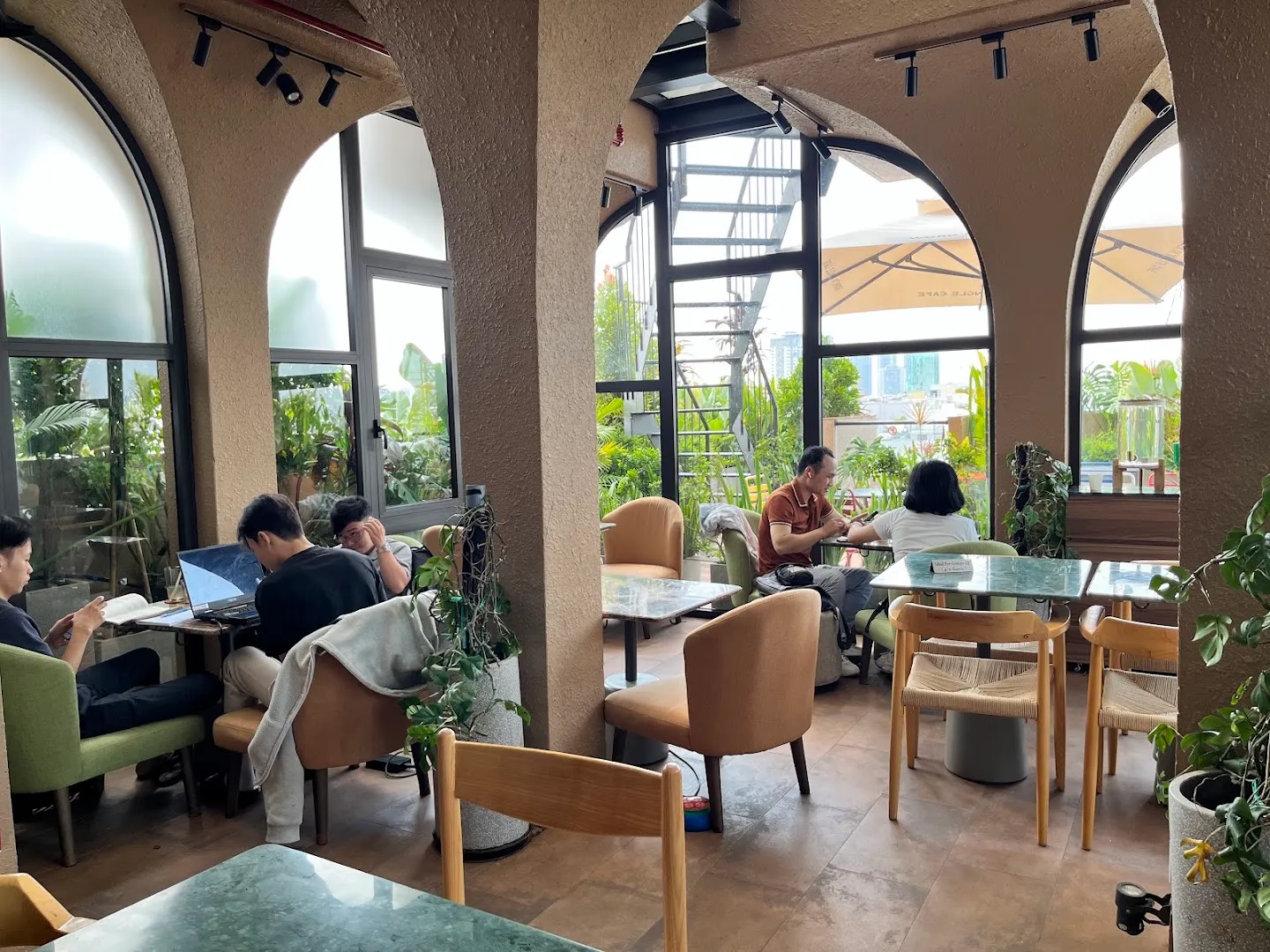 Không gian quán cà phê nhiều cây xanh, bàn ghế thoải mái và ánh sáng tự nhiên.