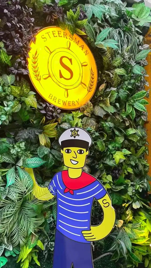 Biển hiệu Steersman Brewery nổi bật giữa mảng tường cây xanh, kèm hình minh họa thủy thủ.