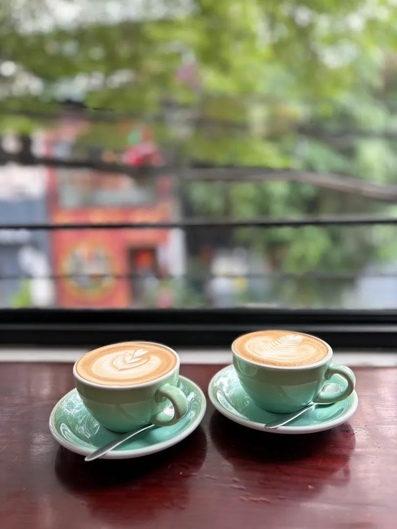 Hai tách cà phê latte với lớp tạo hình trên bàn gỗ cạnh cửa sổ