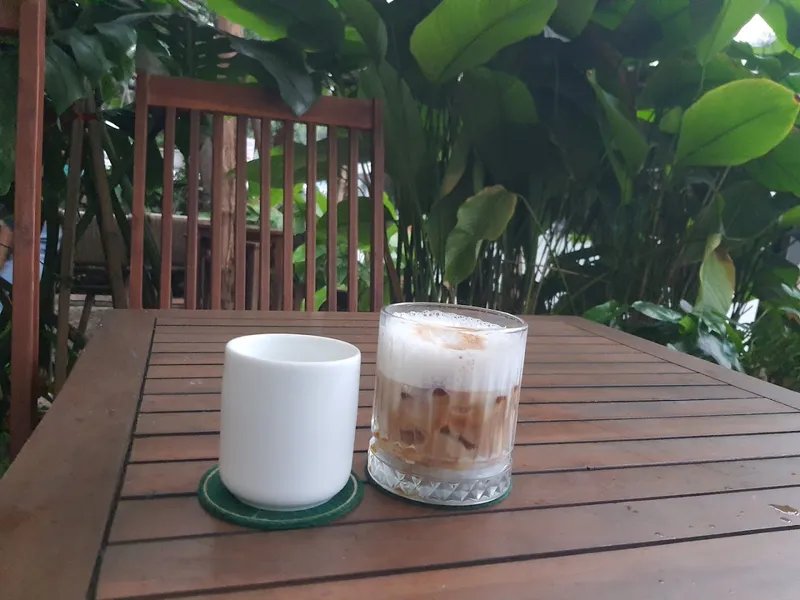 Ly cà phê sữa đá trên bàn gỗ giữa không gian nhiều cây xanh.