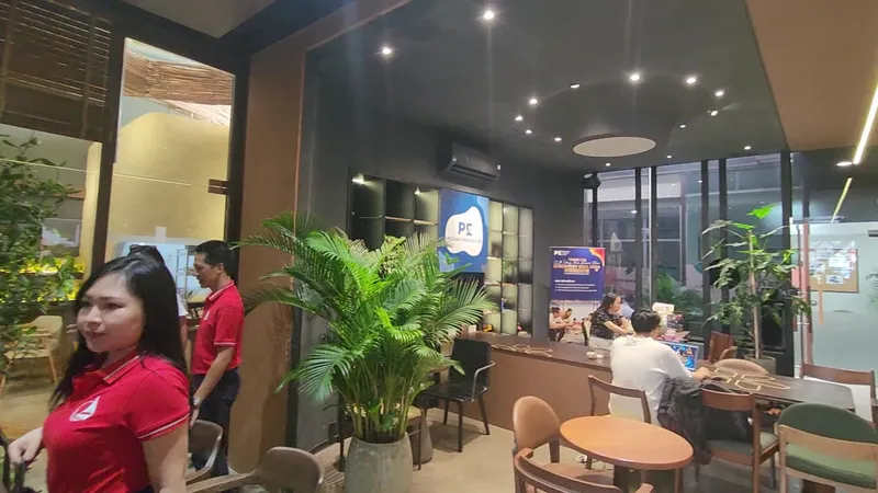 Không gian quán cà phê với bàn ghế, cây xanh và khu vực ngồi bên cửa kính.