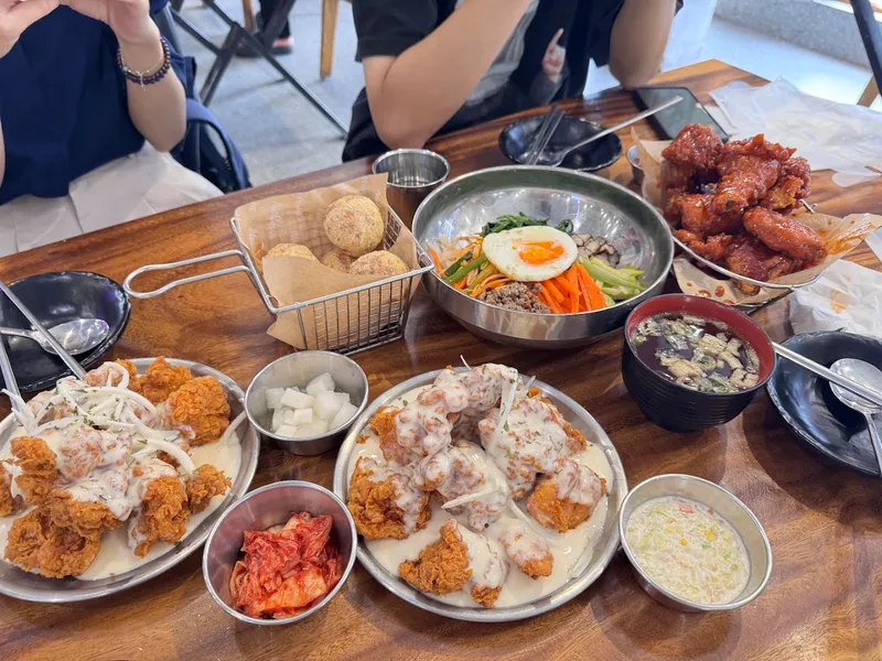 Bàn ăn với gà rán sốt kem, gà sốt cay, cơm trộn bibimbap và các món ăn kèm kiểu Hàn Quốc.