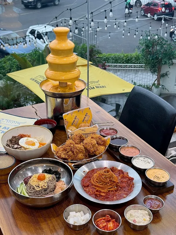 Bàn ăn với gà rán, mì, cơm và nhiều loại sốt, kèm tháp phô mai tan chảy.