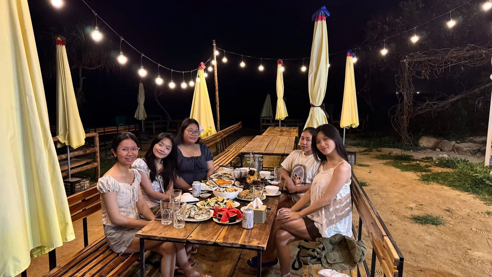 Không gian ngoài trời với bàn gỗ, ô lớn và đèn dây, nơi một nhóm khách đang dùng bữa.