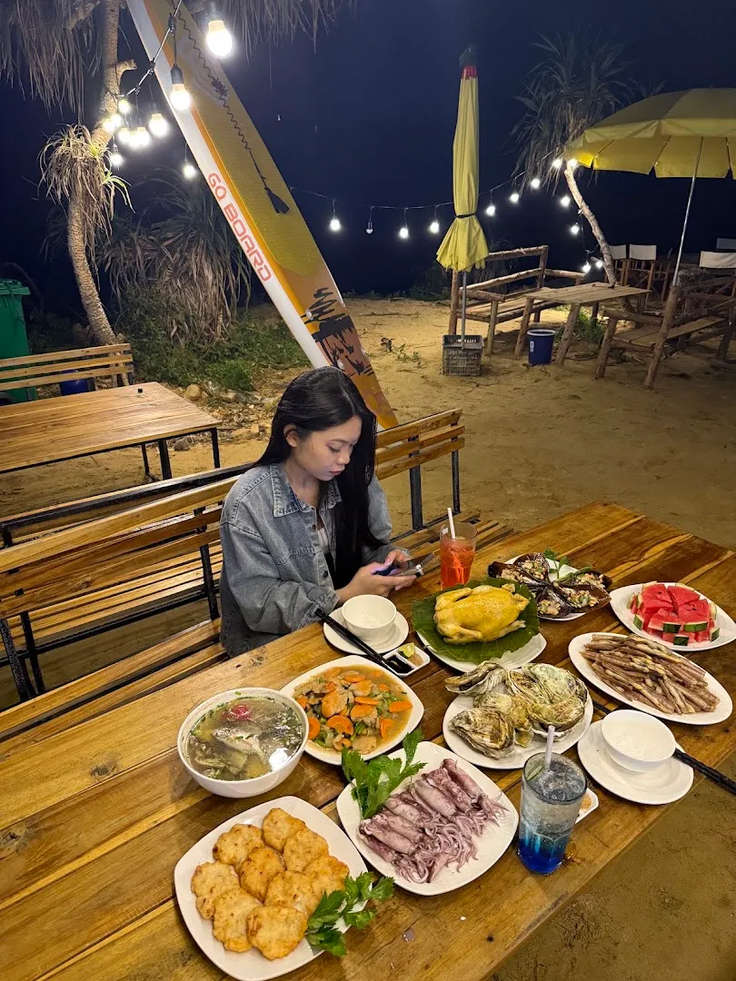 Không gian quán ngoài trời về đêm với bàn ghế gỗ, đèn dây và các món ăn được bày trên bàn.