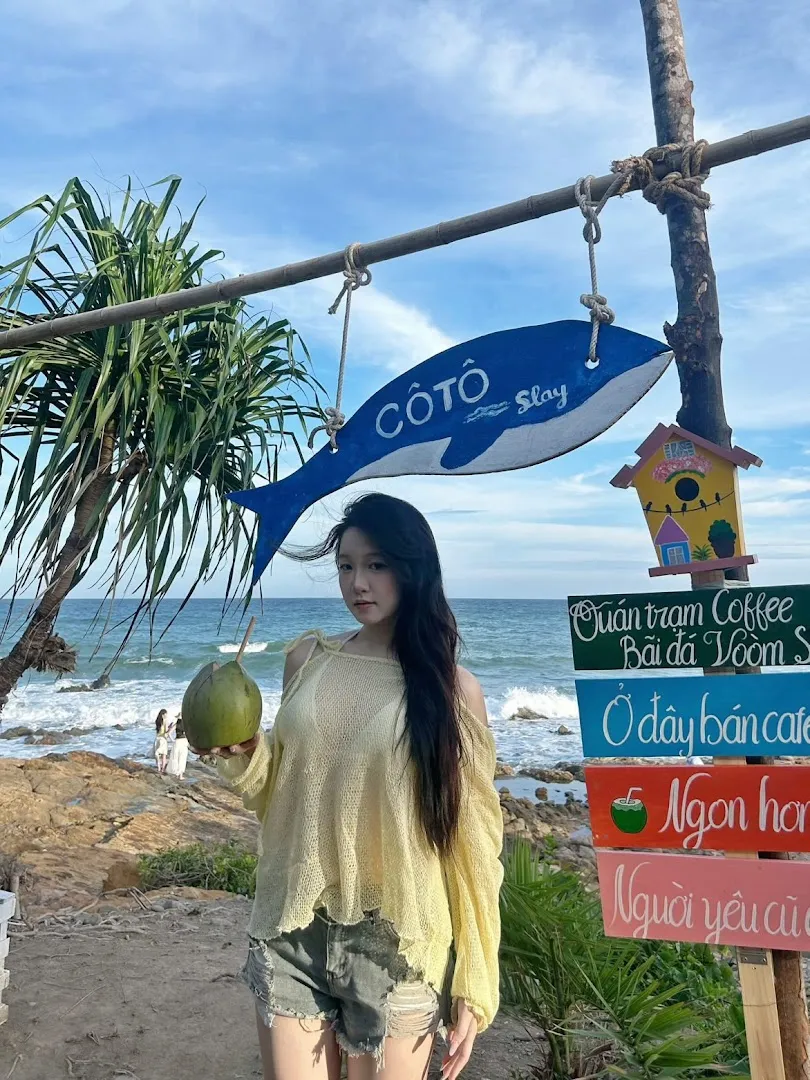 Bảng hiệu Trạm Coffee - Bãi đá Vòm Si bên bờ biển, cùng một du khách cầm quả dừa.