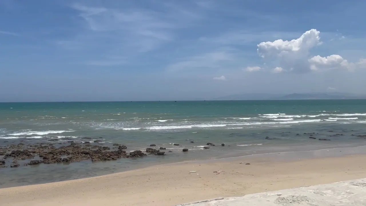 Bãi biển với bờ cát và mặt nước xanh trải dài dưới bầu trời nhiều mây.