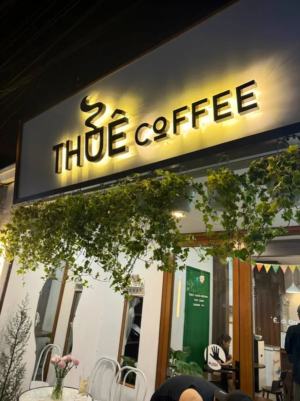 Mặt tiền quán cà phê với bảng hiệu THỎE COFFEE và lối vào phủ cây xanh.