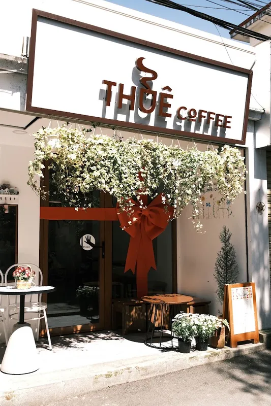 Mặt tiền quán cà phê với biển hiệu “THÓE COFFEE”, cửa ra vào và khu vực bàn ghế phía trước.