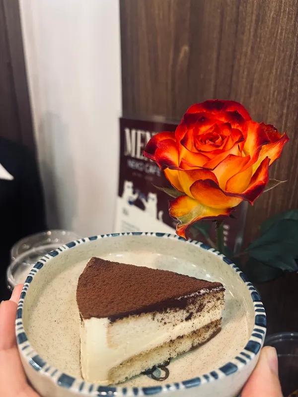 Miếng bánh tiramisu phủ bột cacao trên đĩa, kèm hoa hồng trang trí.