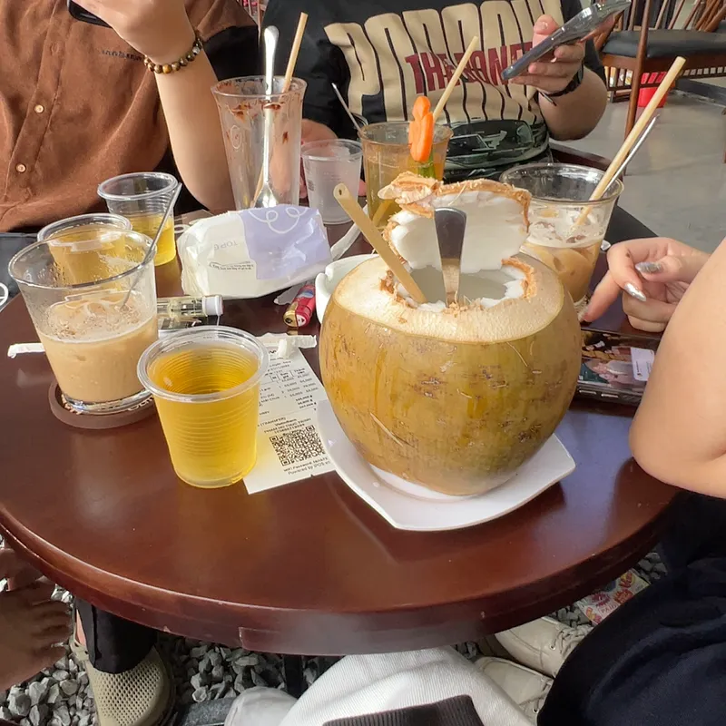 Bàn cà phê với các loại đồ uống, nổi bật là trái dừa phục vụ cùng đá và ống hút.