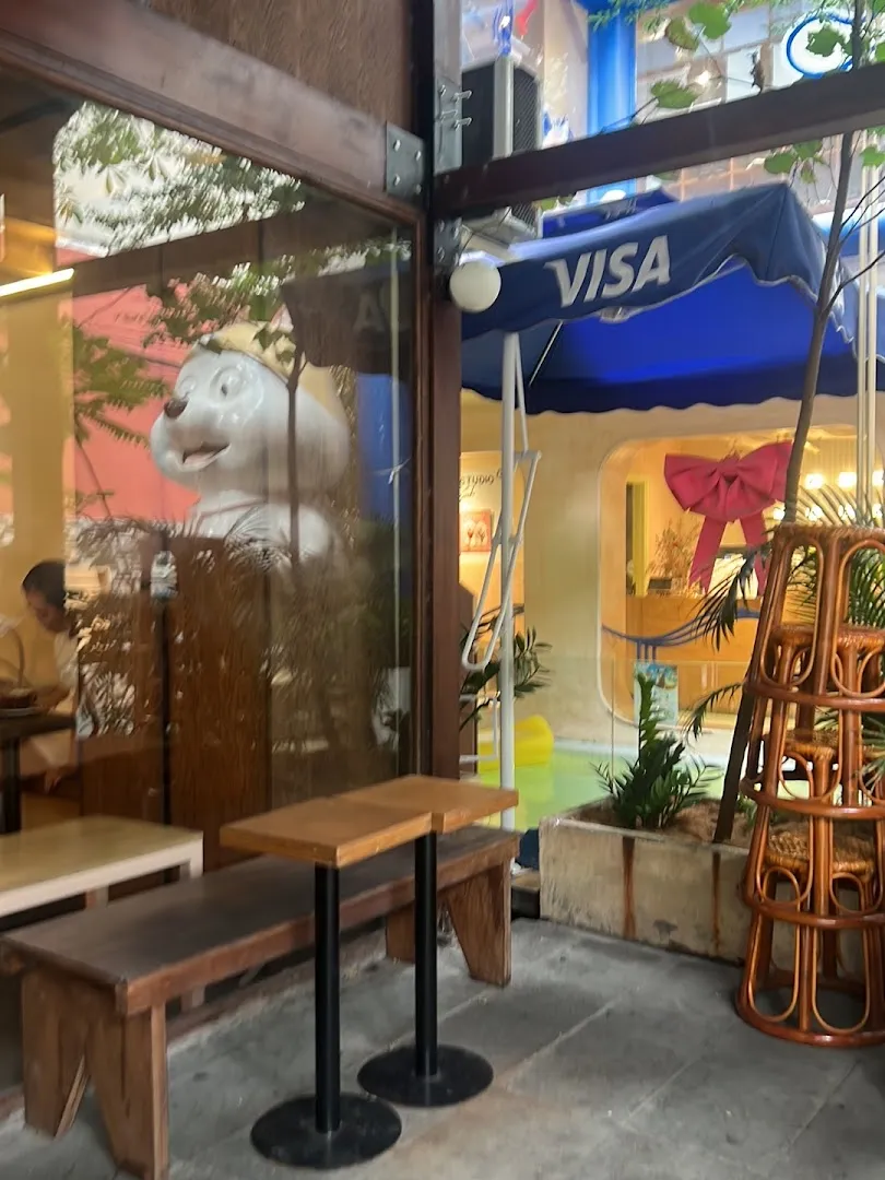 Không gian quán với bàn ghế gỗ, cây xanh và lối vào mở.